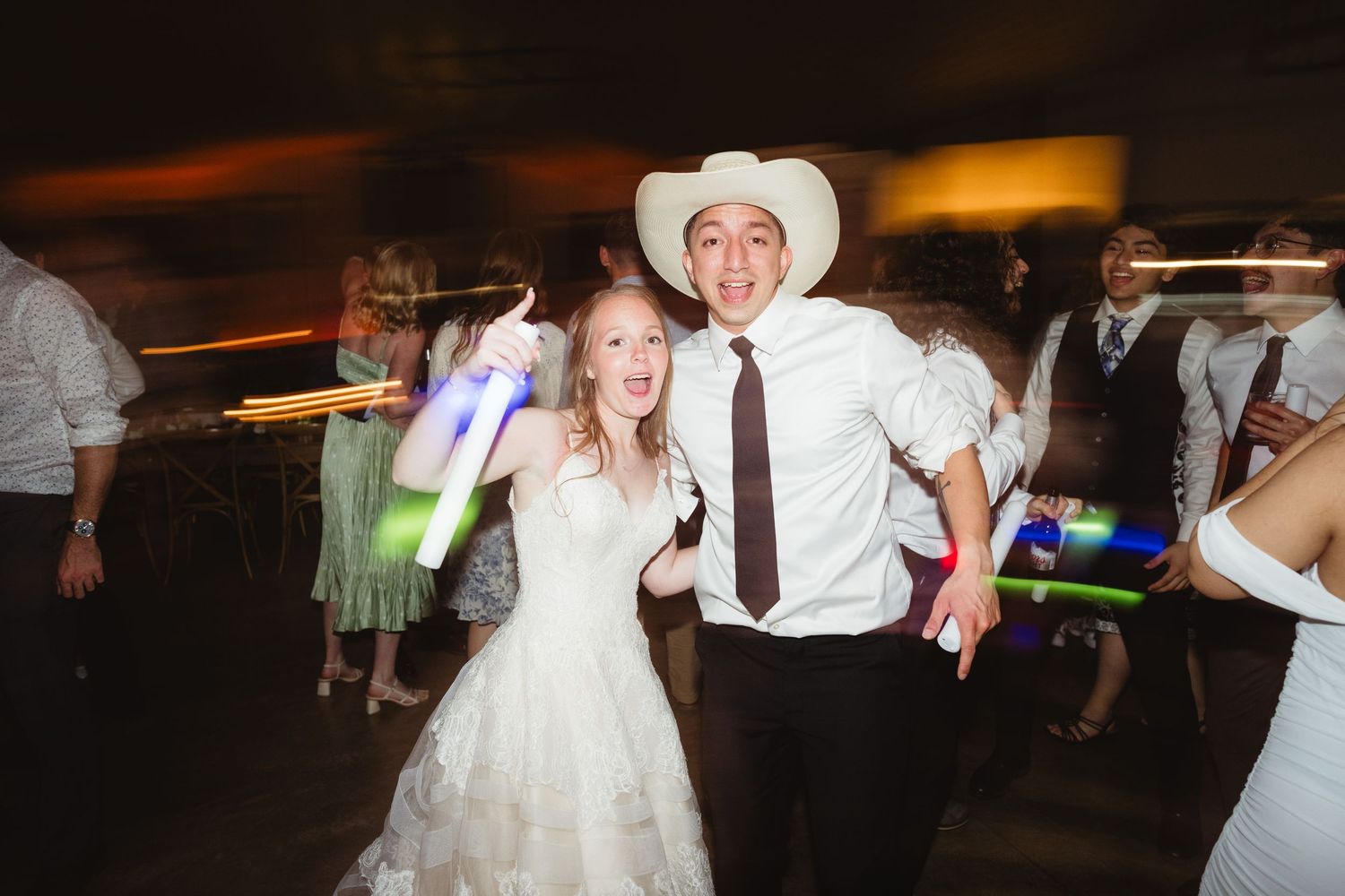 Wedding couple dances together at reception with guests making light trails in dark room.