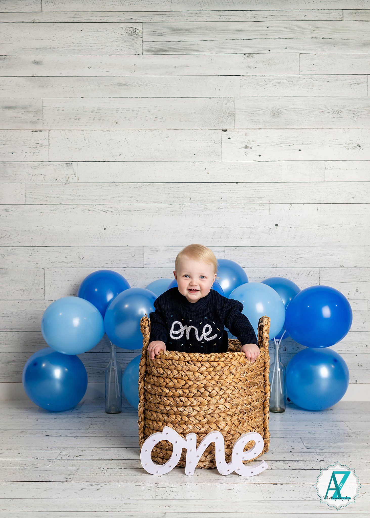 A baby in a black 'one' shirt sits in a woven basket surrounded by blue balloons against a white wood wall.