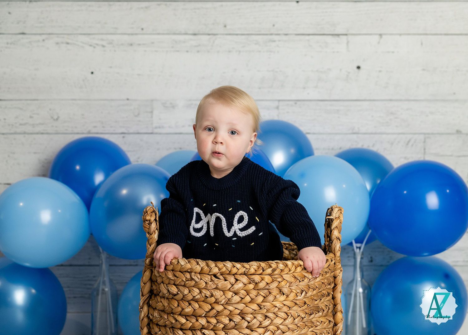 A child in a black 'one' shirt sits in a woven basket with blue balloons in the background against a white wall.