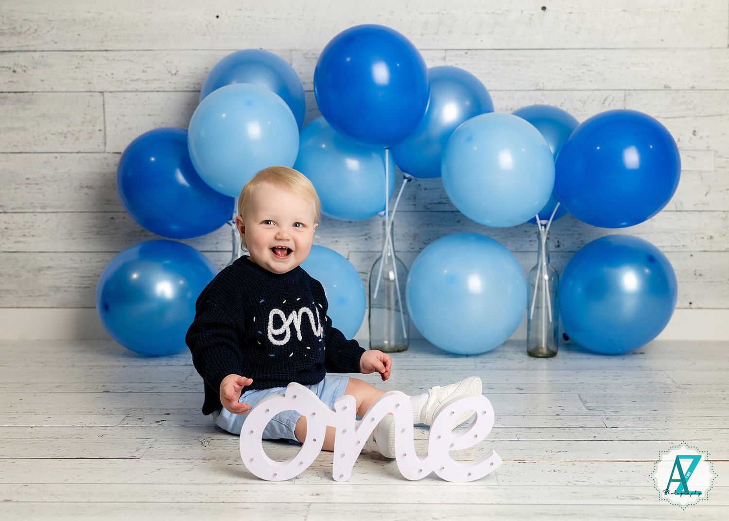 First birthday celebration with blue balloons and white 'one' lettering against white wooden wall background.