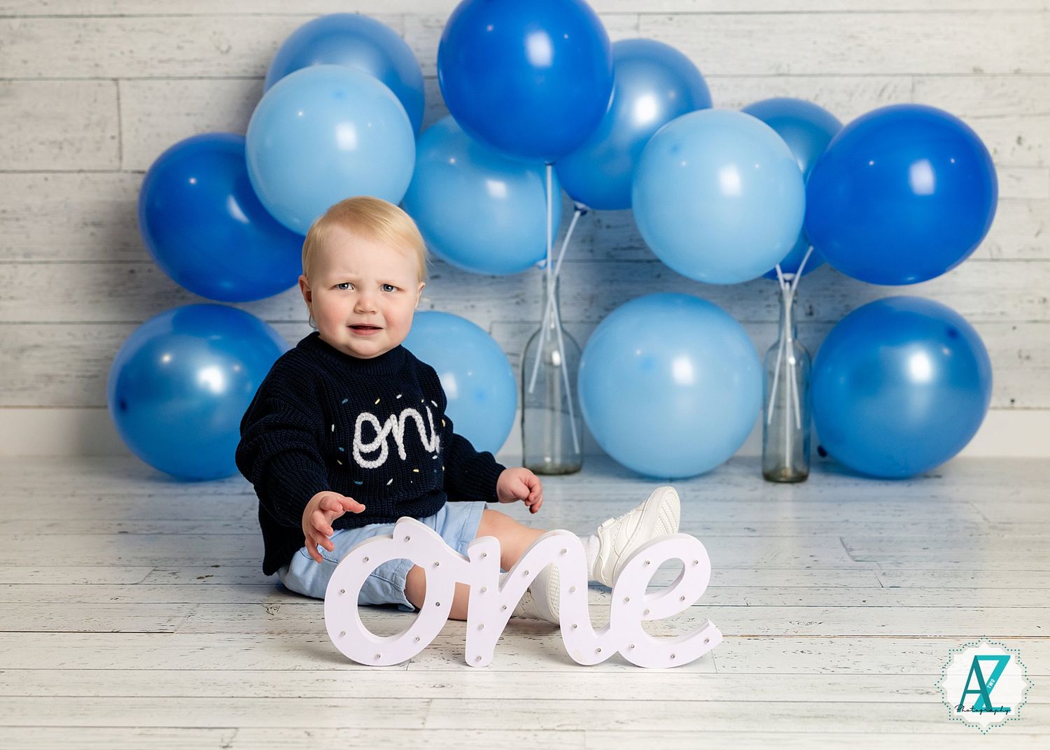 First birthday photo setup with light and dark blue balloons arranged in an arch with white 'one' letters on wooden floor.