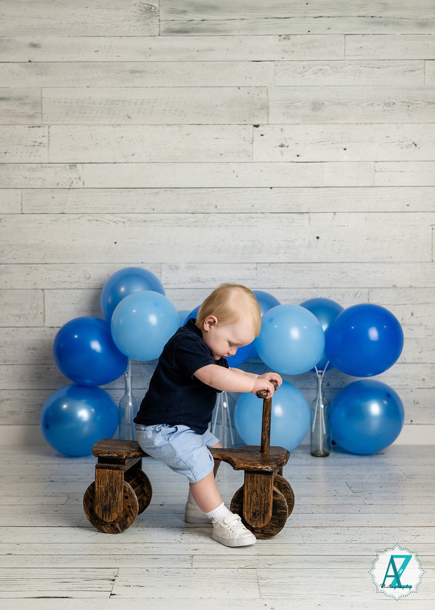 A small child in black shirt sits on a vintage wooden balance bike against a white wall with blue balloons.