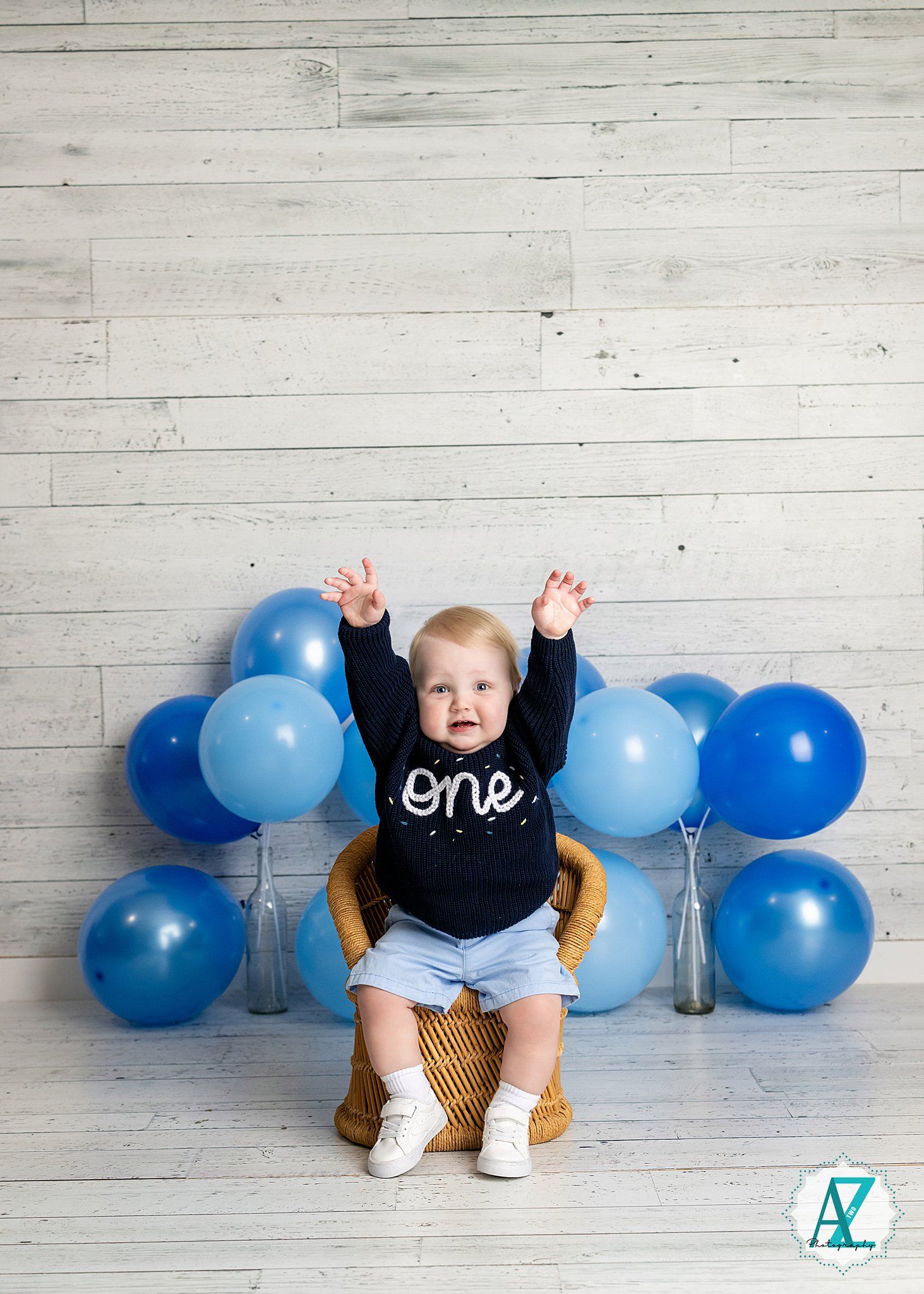 A small child in a 'one' shirt sits on a chair with blue balloons decorating the background.