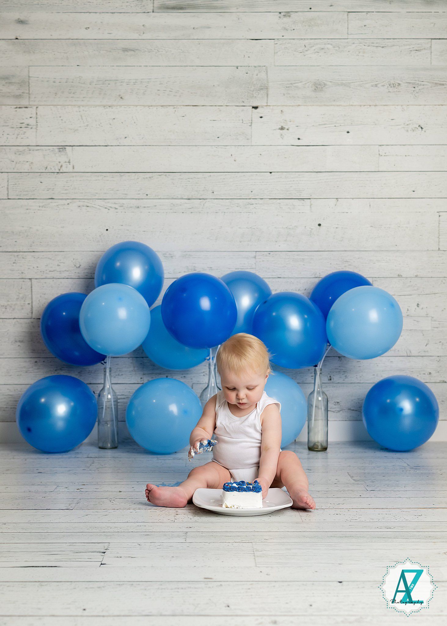Baby in white outfit sits with birthday cake surrounded by blue balloons against rustic wood wall backdrop.