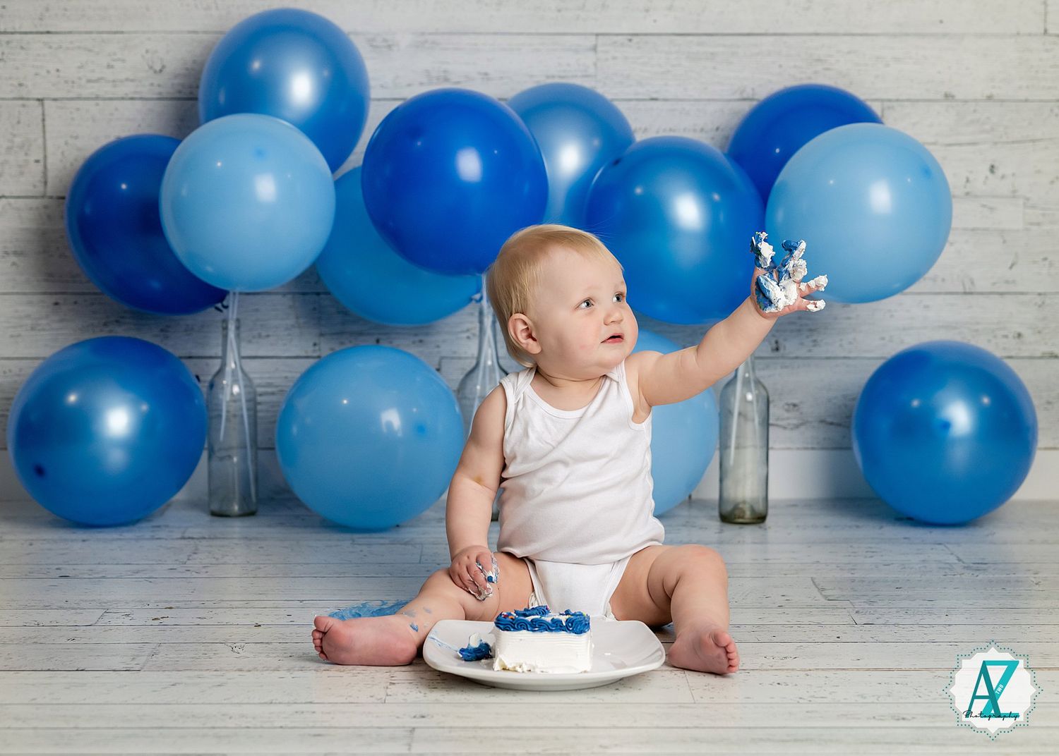 Baby in white onesie sits with cake against backdrop of blue balloons during first birthday photoshoot.