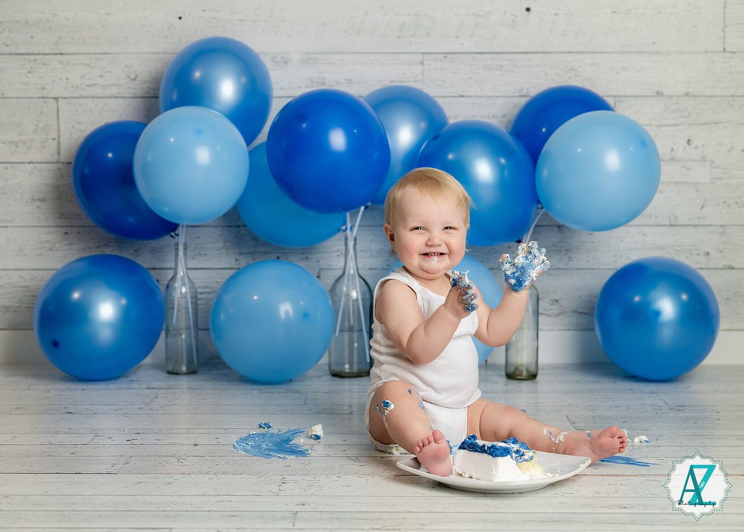 A small baby plays with cake and decorations while surrounded by blue balloons against a wooden wall.