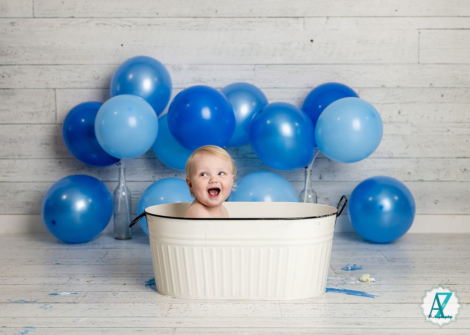 A small baby sits in a white tub with joyful expression surrounded by blue balloons against a wooden wall backdrop.