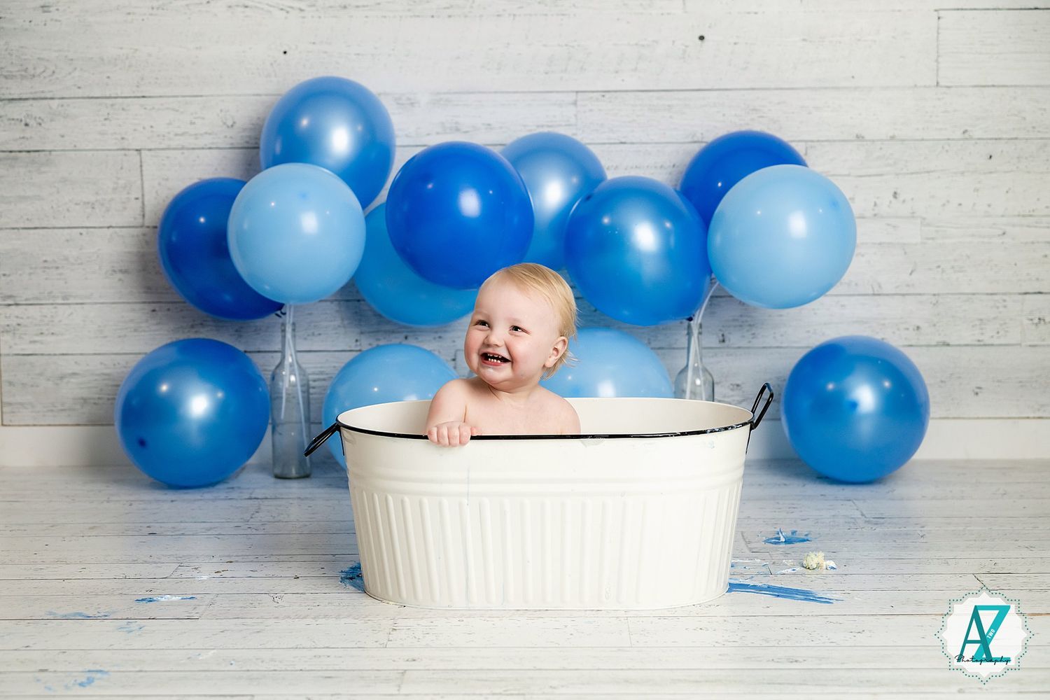 Happy baby sitting in white wash basin with blue balloons arranged behind against wooden wall background.