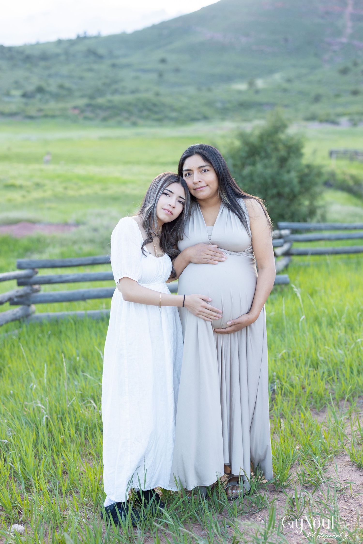 Two people in flowing white and beige dresses pose together in a green mountain meadow with a rustic fence.