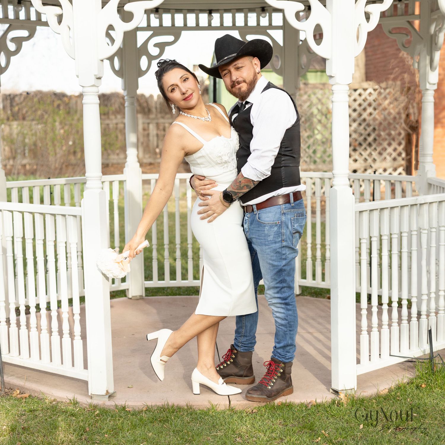 A couple in western-style wedding attire embraces in a white gazebo on a sunny day.