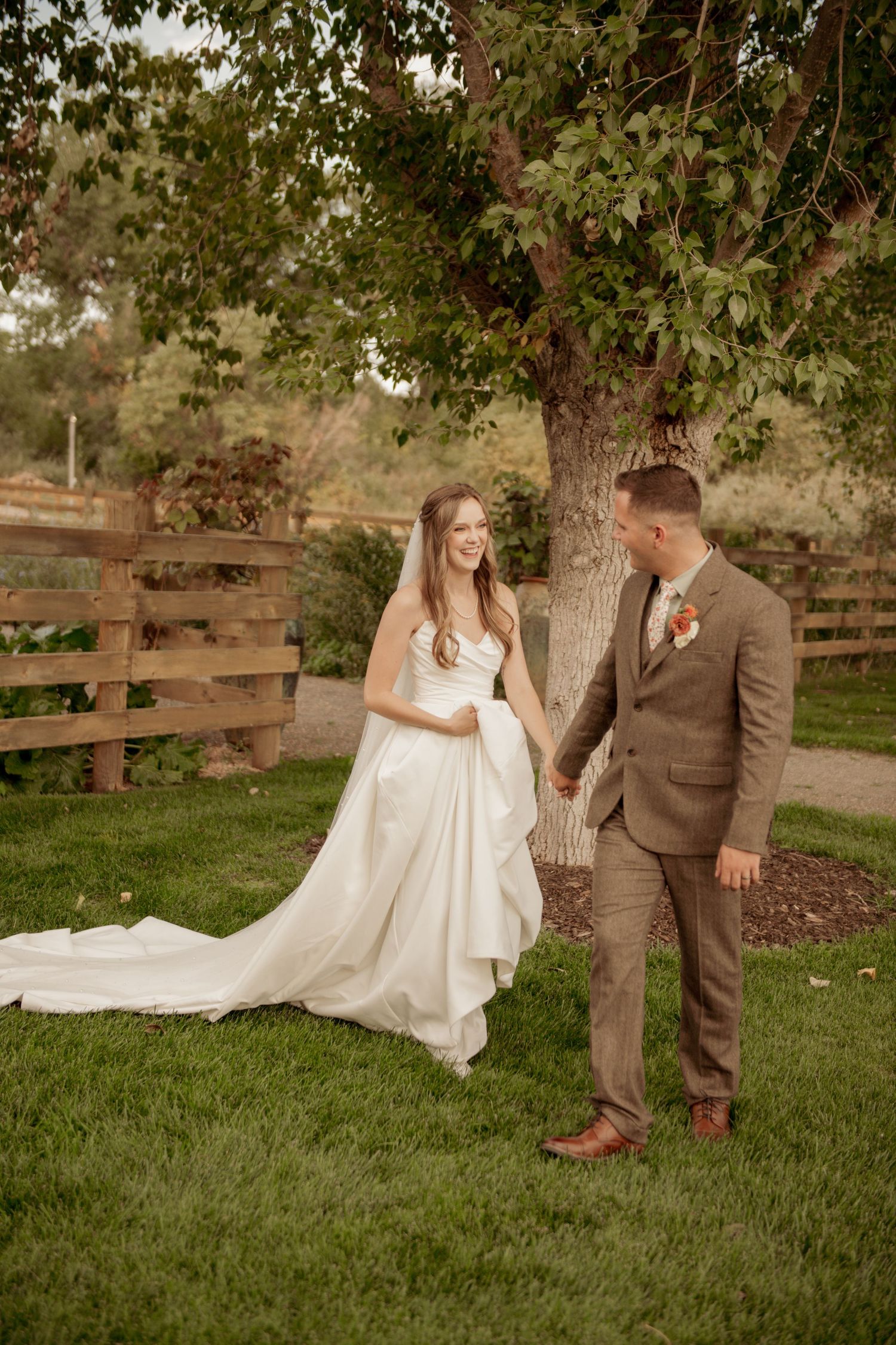 A couple poses for a wedding photo on a lawn with a rustic wooden fence and tree in the background.