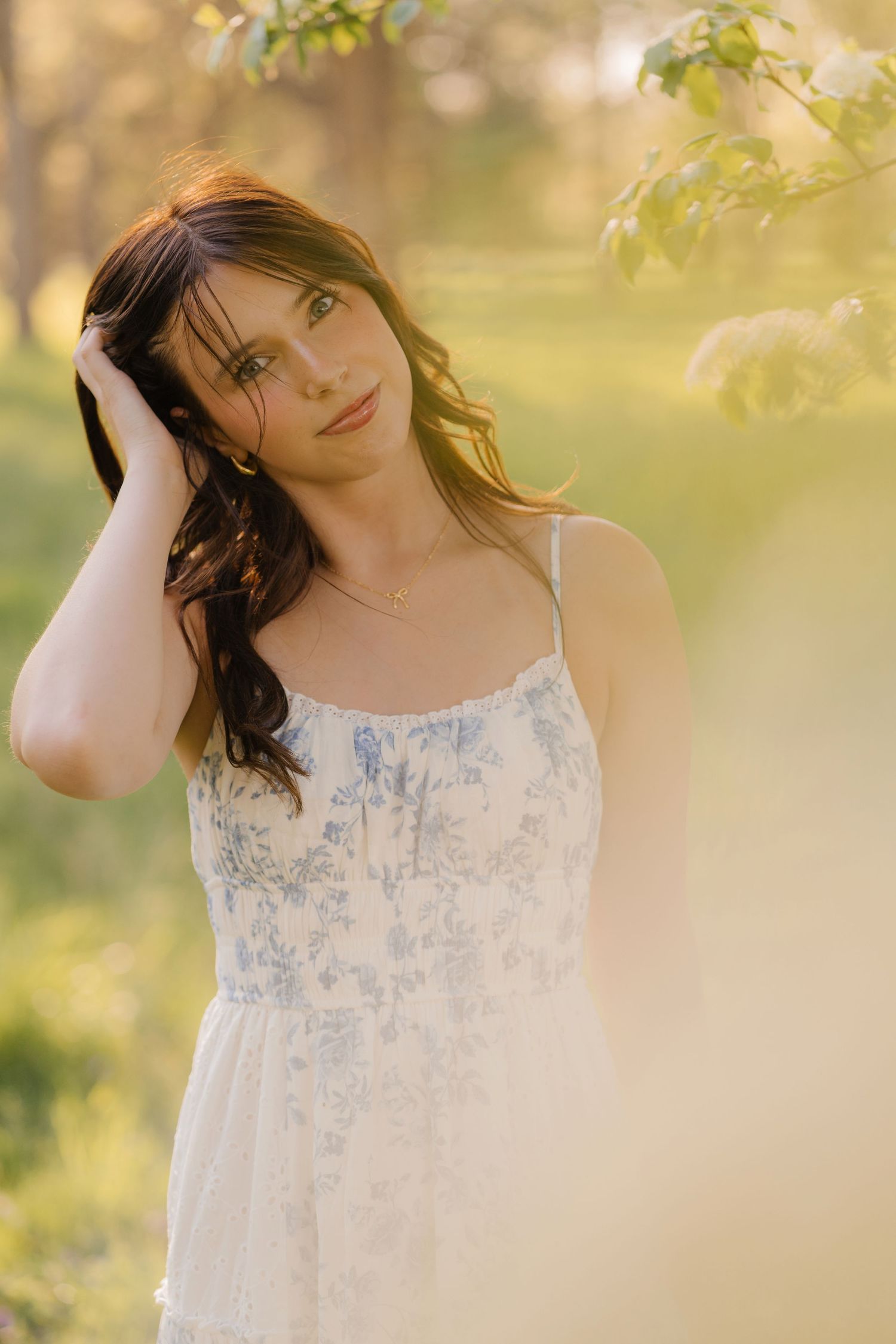 A portrait of someone in a blue and white floral sundress posing near a tree trunk with ivy on a sunny day.