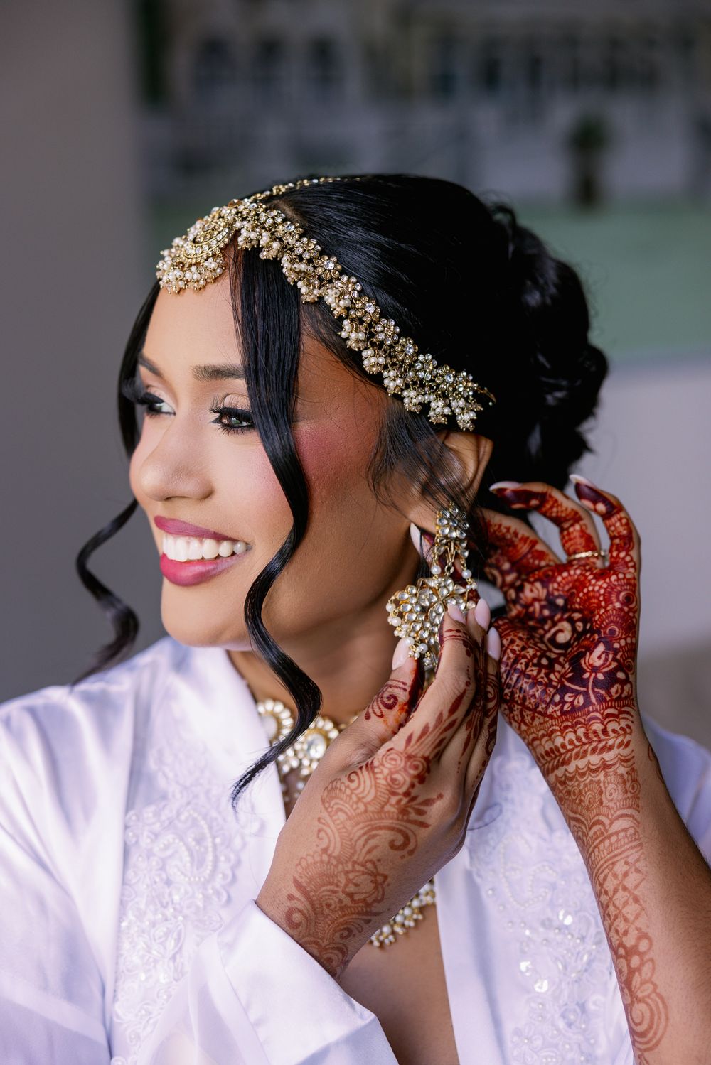Bride wearing gold headpiece and white lace robe puts on earrings with traditional henna-adorned hands.
