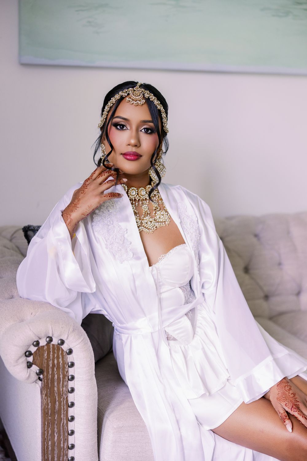 Elegant bridal portrait with white silk robe, ornate gold jewelry and headpiece sitting on tufted couch.