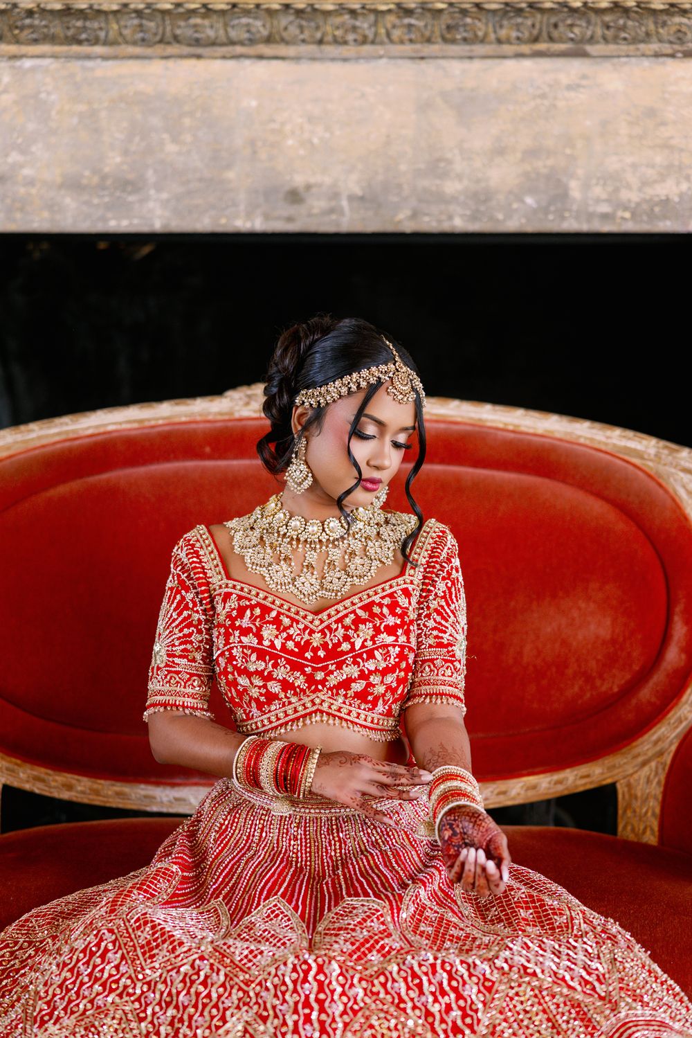 Traditional Indian bride in ornate red and gold lehenga sits elegantly on antique red sofa.