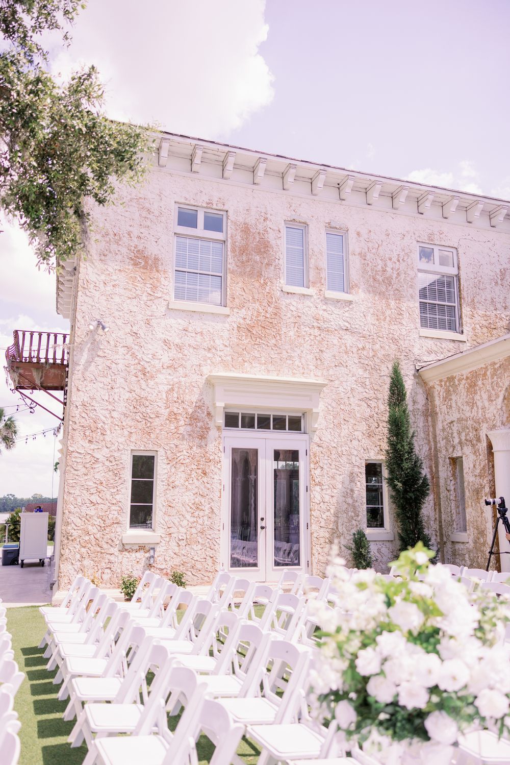 White chairs arranged for an outdoor wedding ceremony in front of a rustic stone building with white floral decorations.
