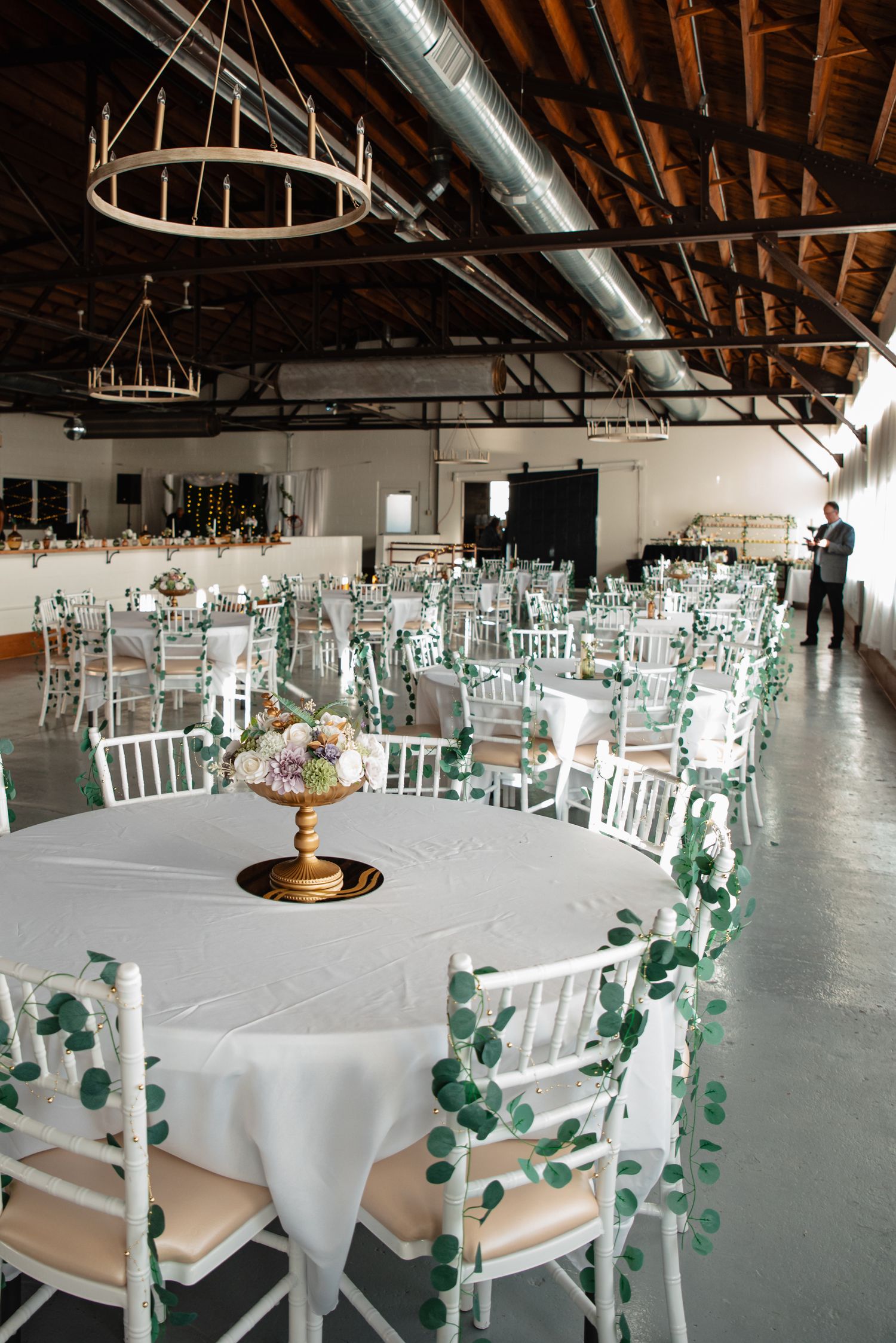 Wedding reception venue with round white tables, mint green chairs draped in eucalyptus garlands, and rustic chandeliers overhead.