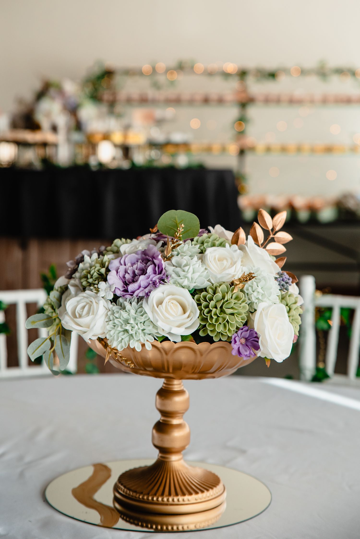 Elegant flower arrangement featuring white roses, purple blooms and succulents in a copper pedestal bowl on white tablecloth.