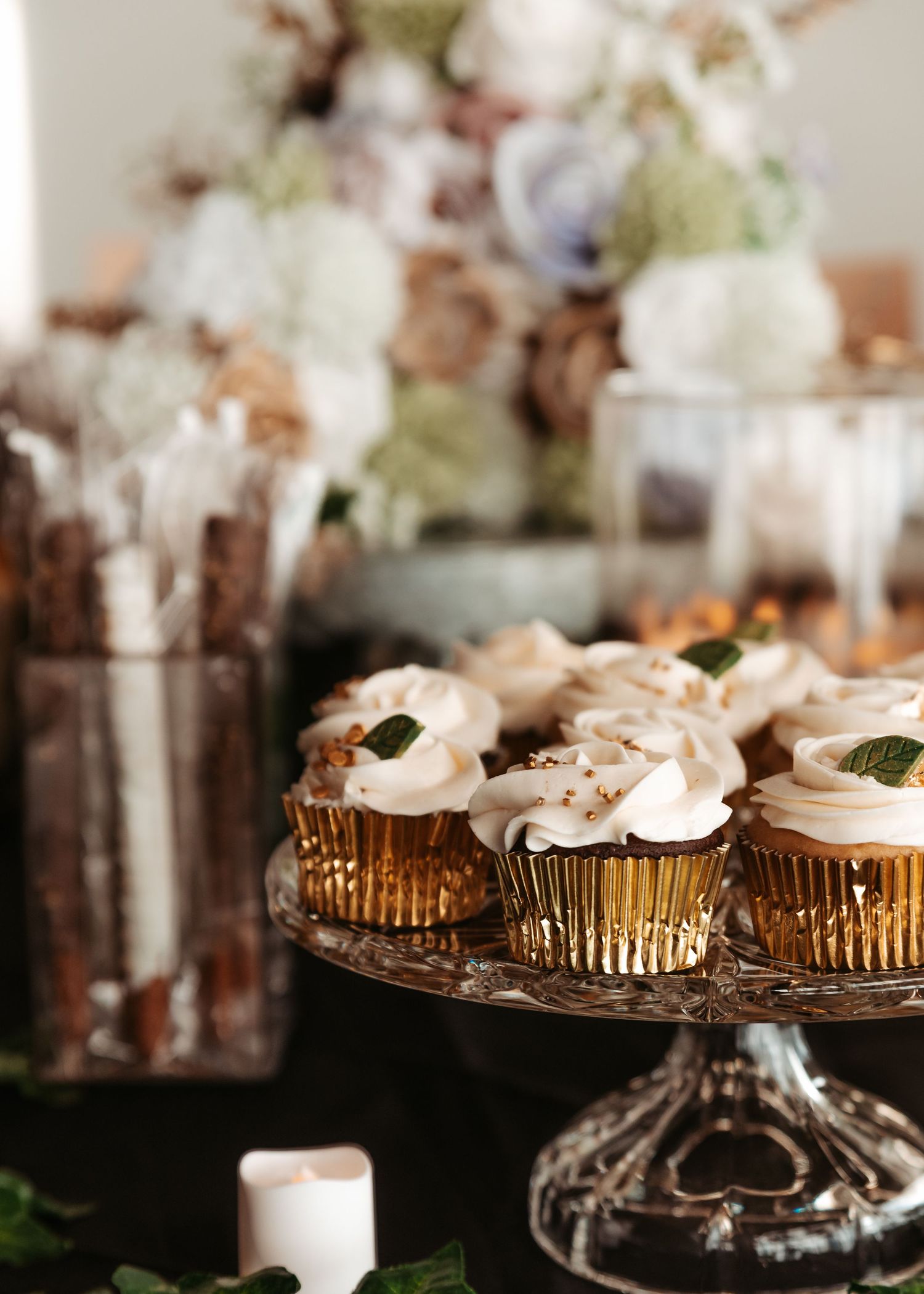 Elegant gold foil cupcakes arranged on a crystal cake stand with soft floral decorations and candlelight in background.