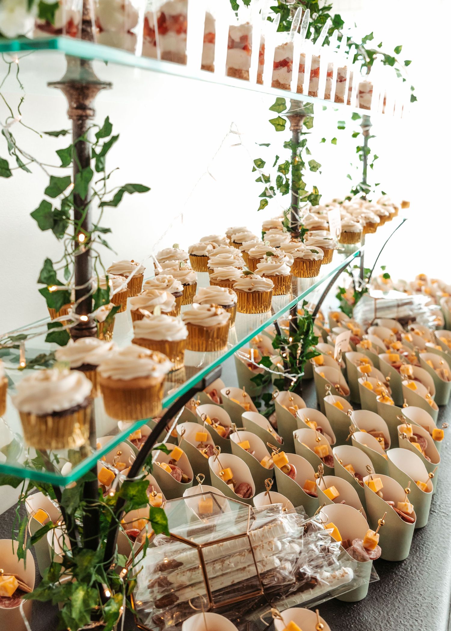 Elegant food display featuring rows of dessert shooters and appetizer bites arranged on glass shelves with green foliage accents.