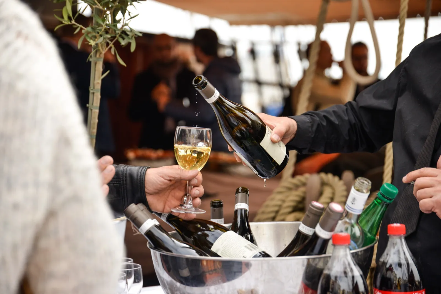 Un serveur verse un verre de vin blanc dans une ambiance de réception, avec bouteilles et convives en arrière-plan.