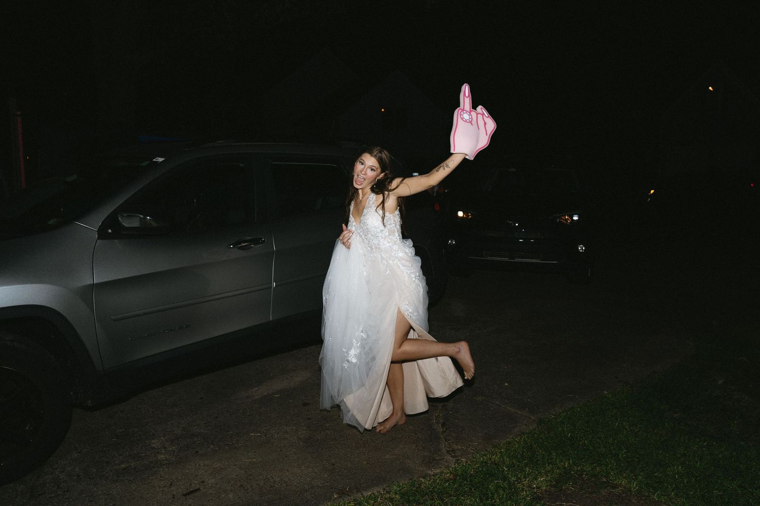 A bride in a flowing white dress holds up a bottle while standing next to a car at night.