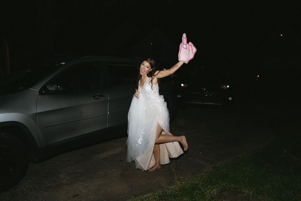A bride in a flowing white dress holds up a bottle while standing next to a car at night.