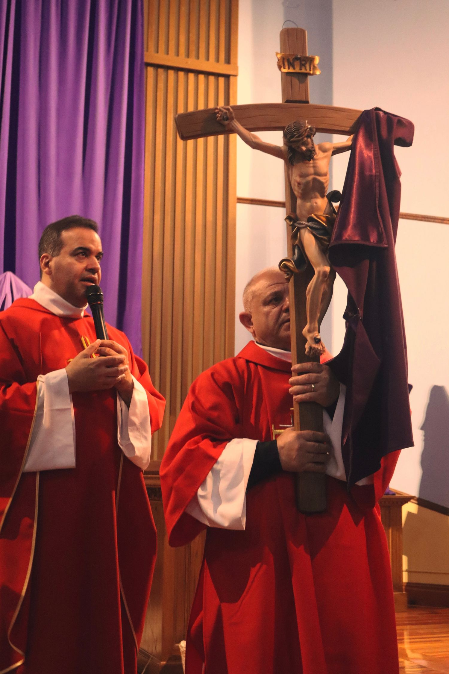 Two clergy members in red vestments conduct Good Friday service with a crucifix in a church interior.