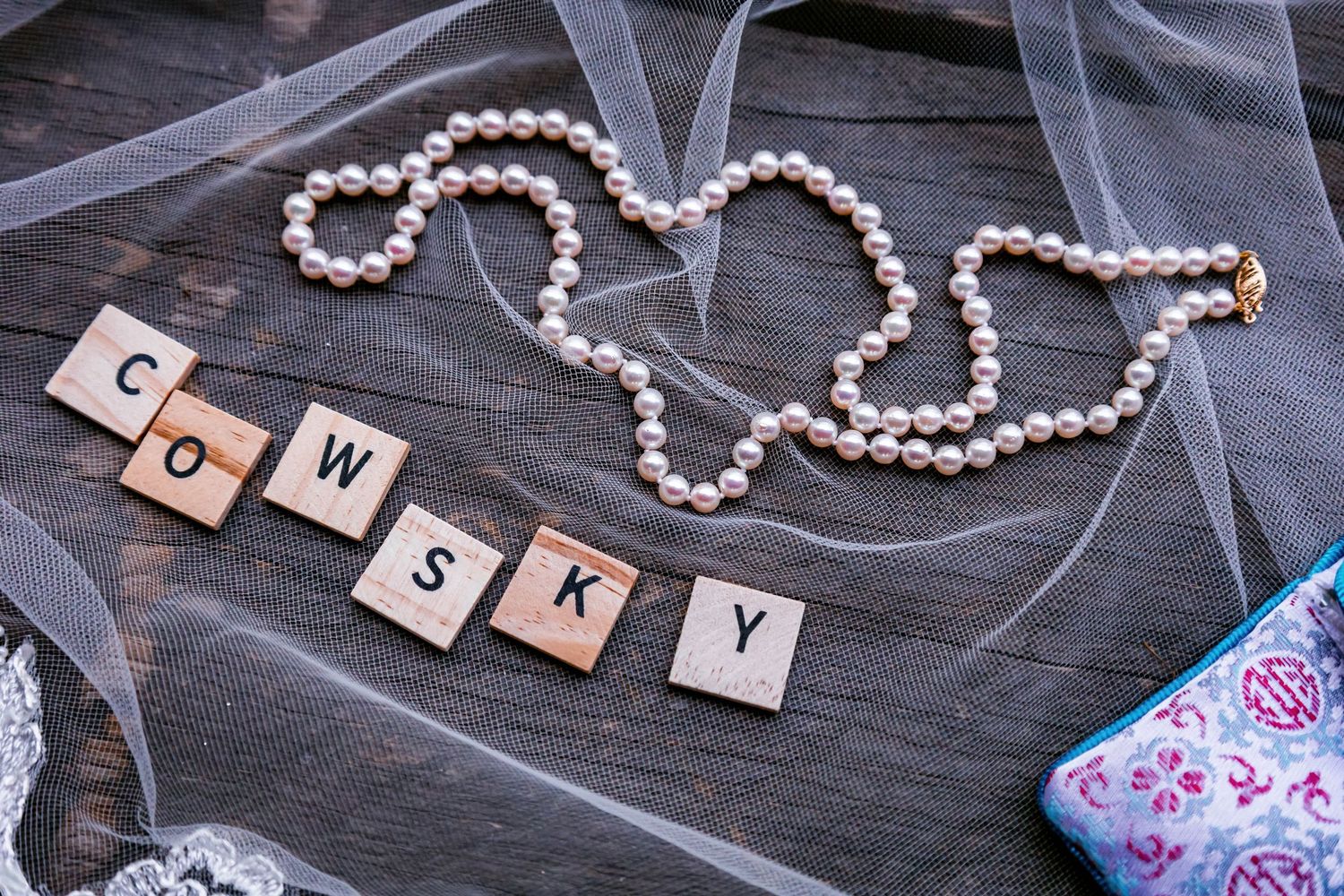 Pearl necklace and scrabble tiles spelling 'Cowsky' arranged with tulle on dark wood.