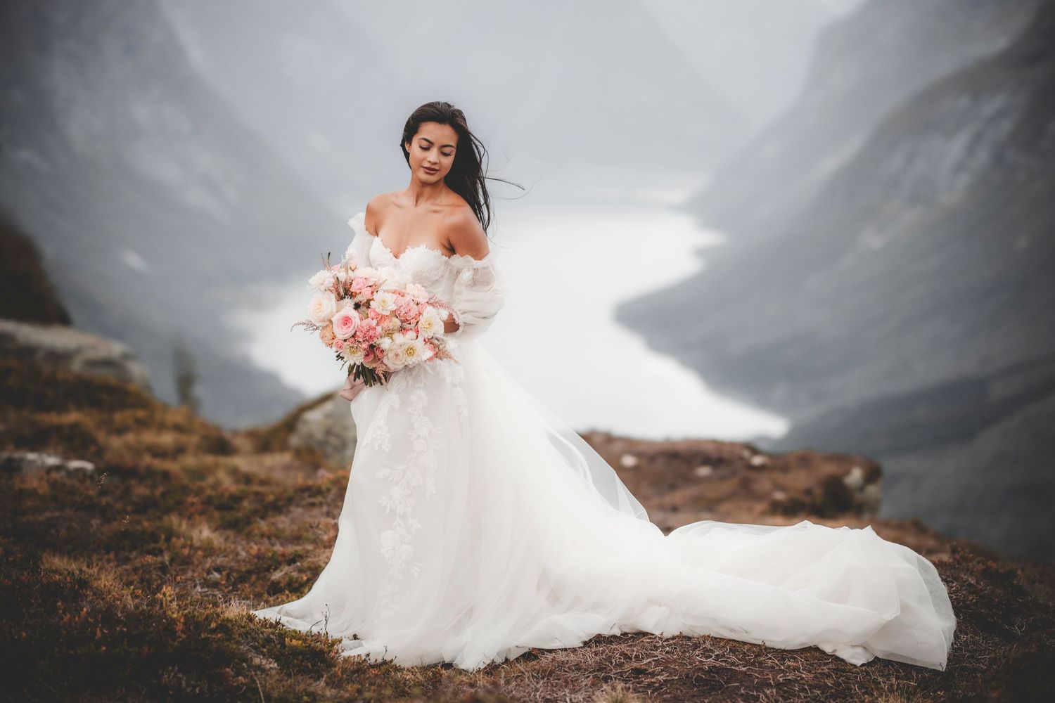A bride in a flowing white gown holds a pink bouquet while standing on a misty mountain ridge in dramatic scenery.