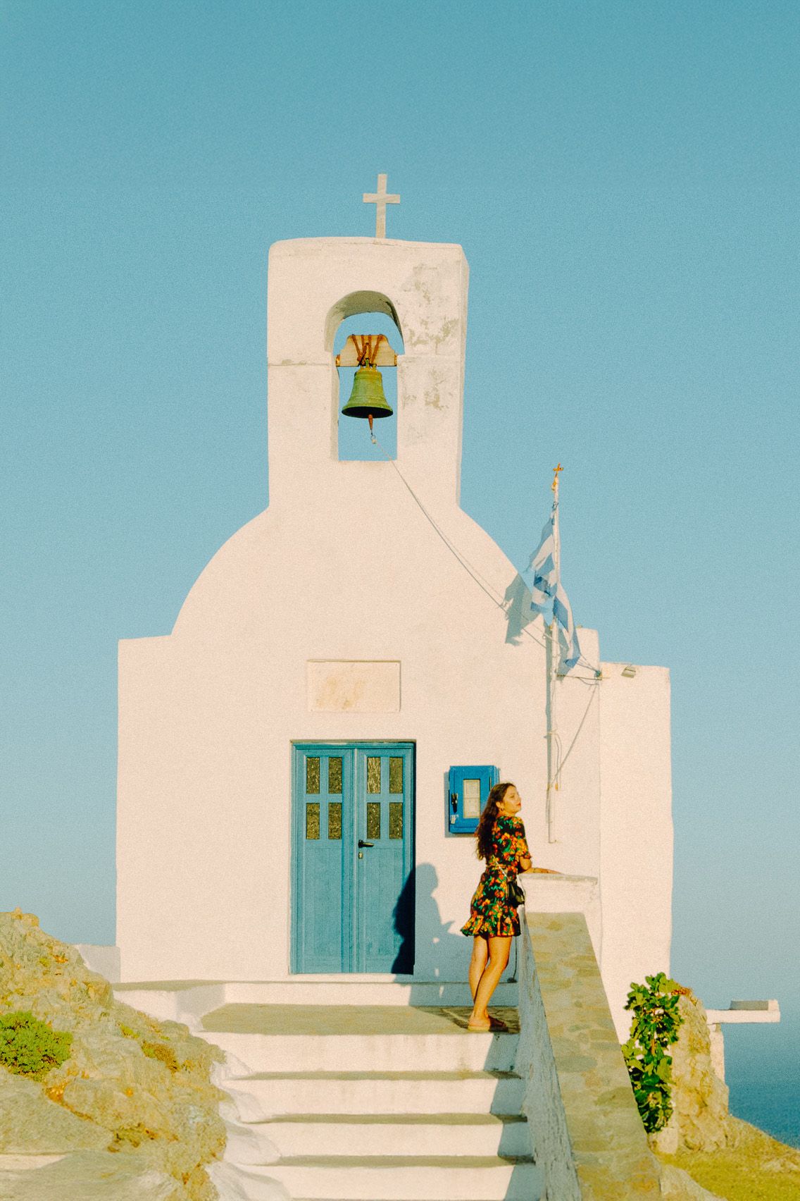 Woman standing on steps of white chapel under blue sky, evoking serene Aegean island travel atmosphere