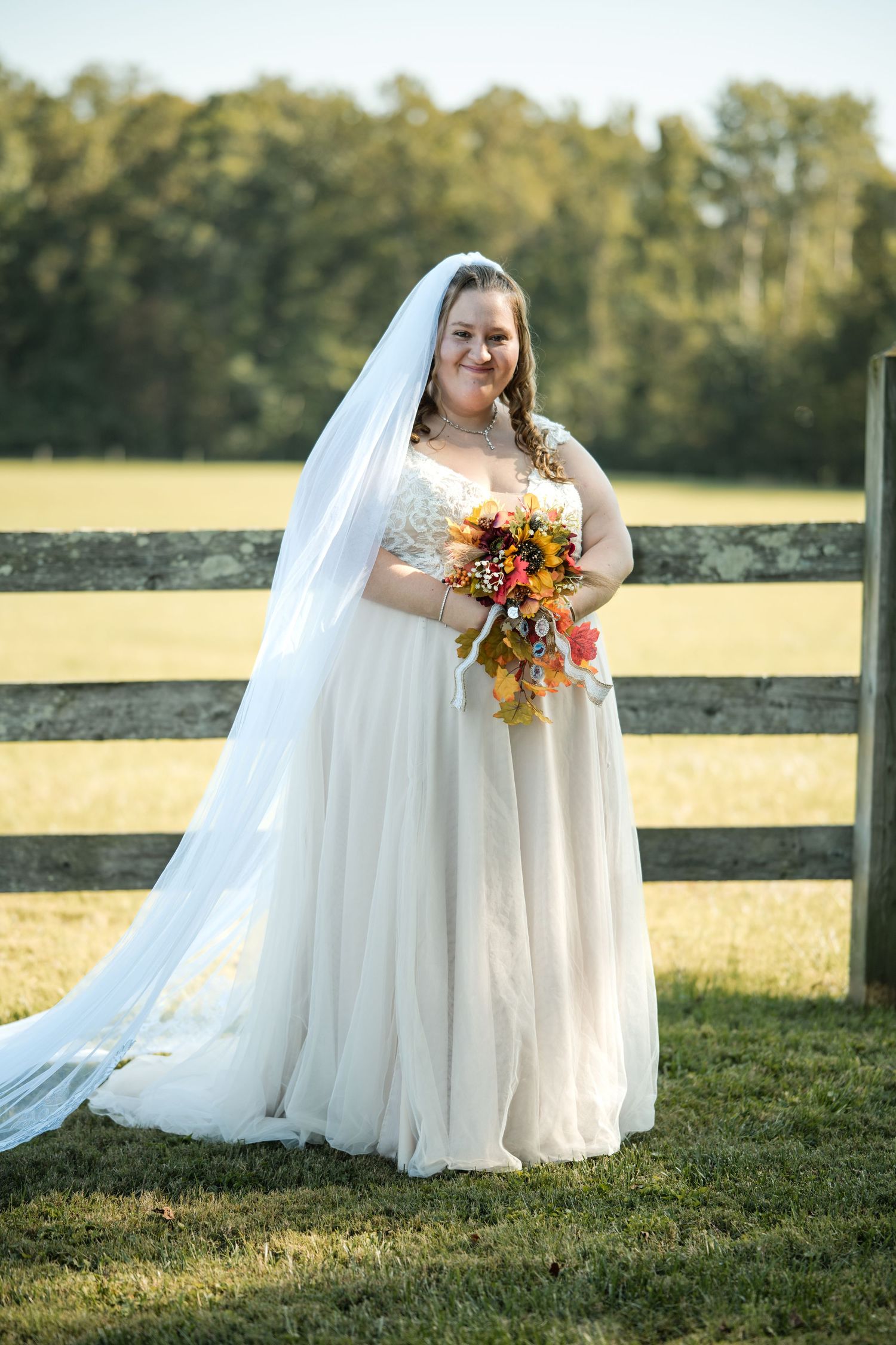 Bride in flowing white dress holds autumn bouquet while posing by wooden fence.