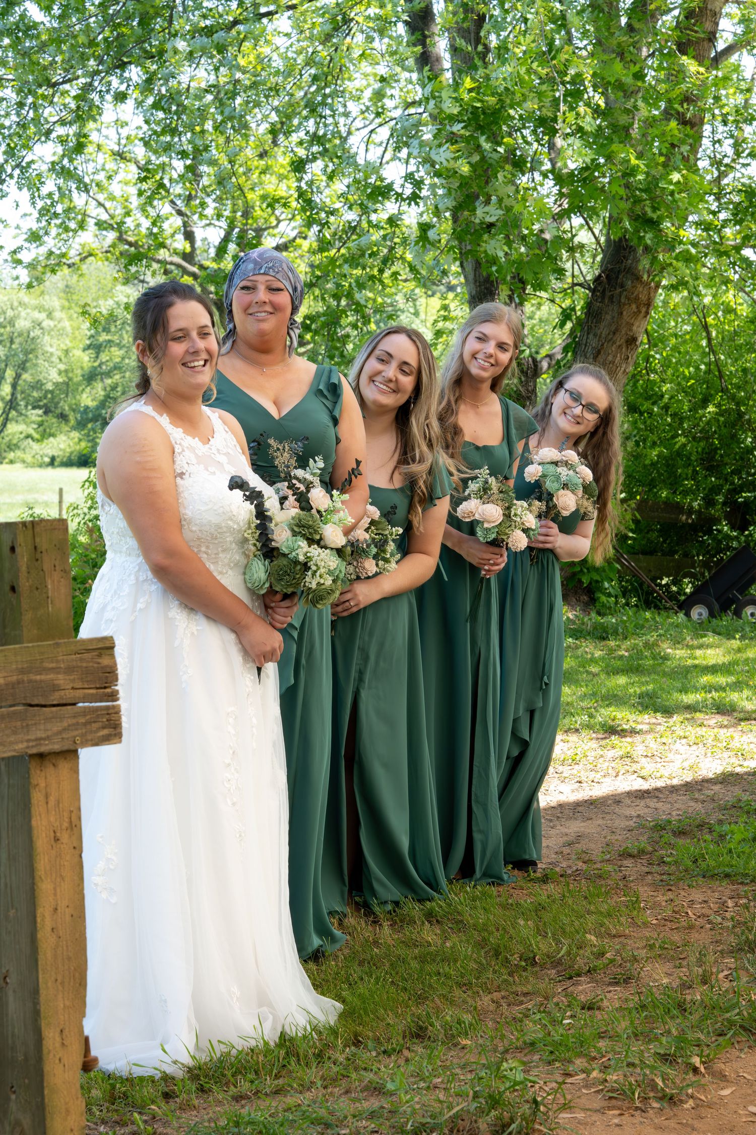 Wedding party in forest green bridesmaid dresses and white bridal gown pose together on wooded outdoor path.