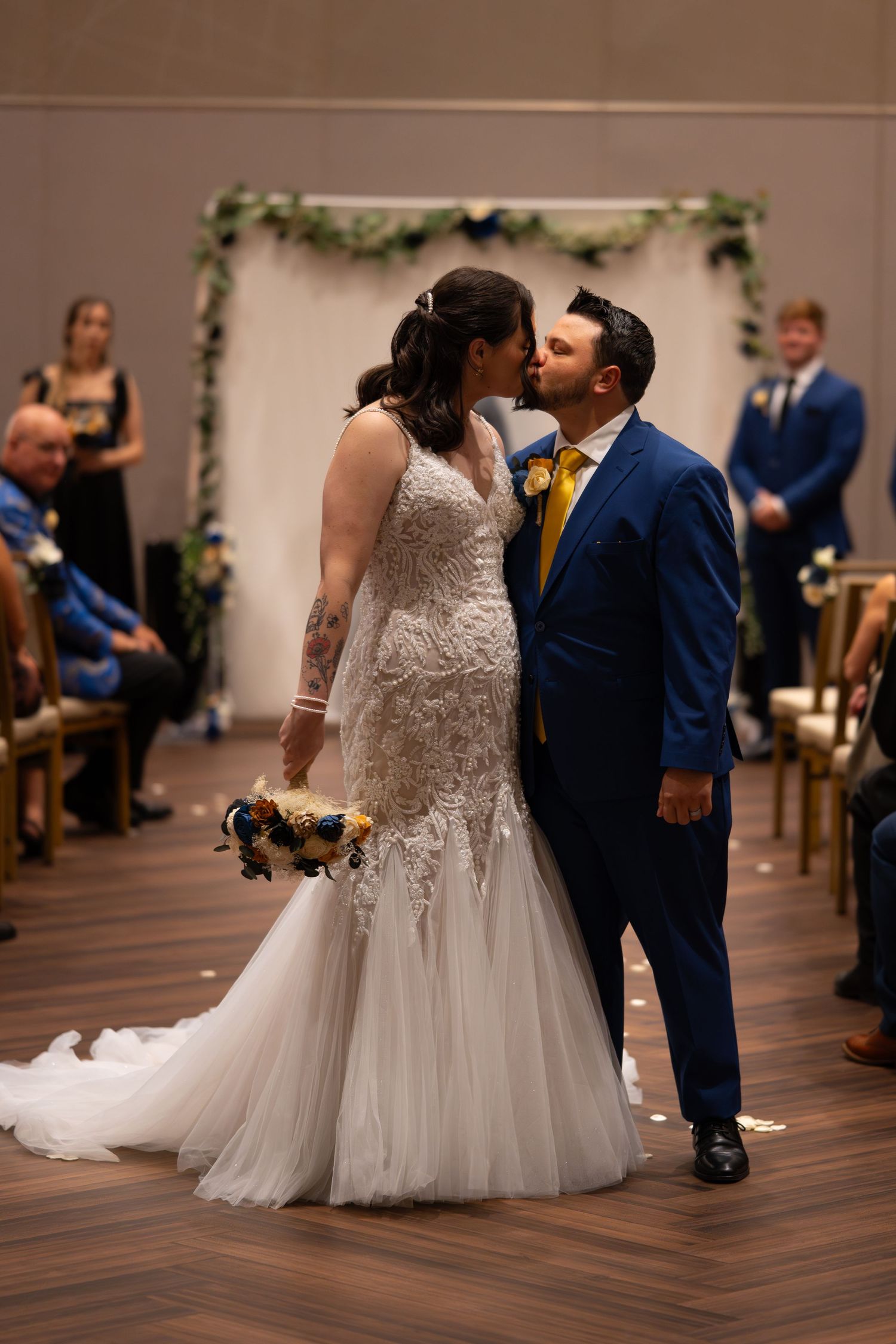 Wedding couple shares their first kiss during indoor ceremony with floral arch backdrop.