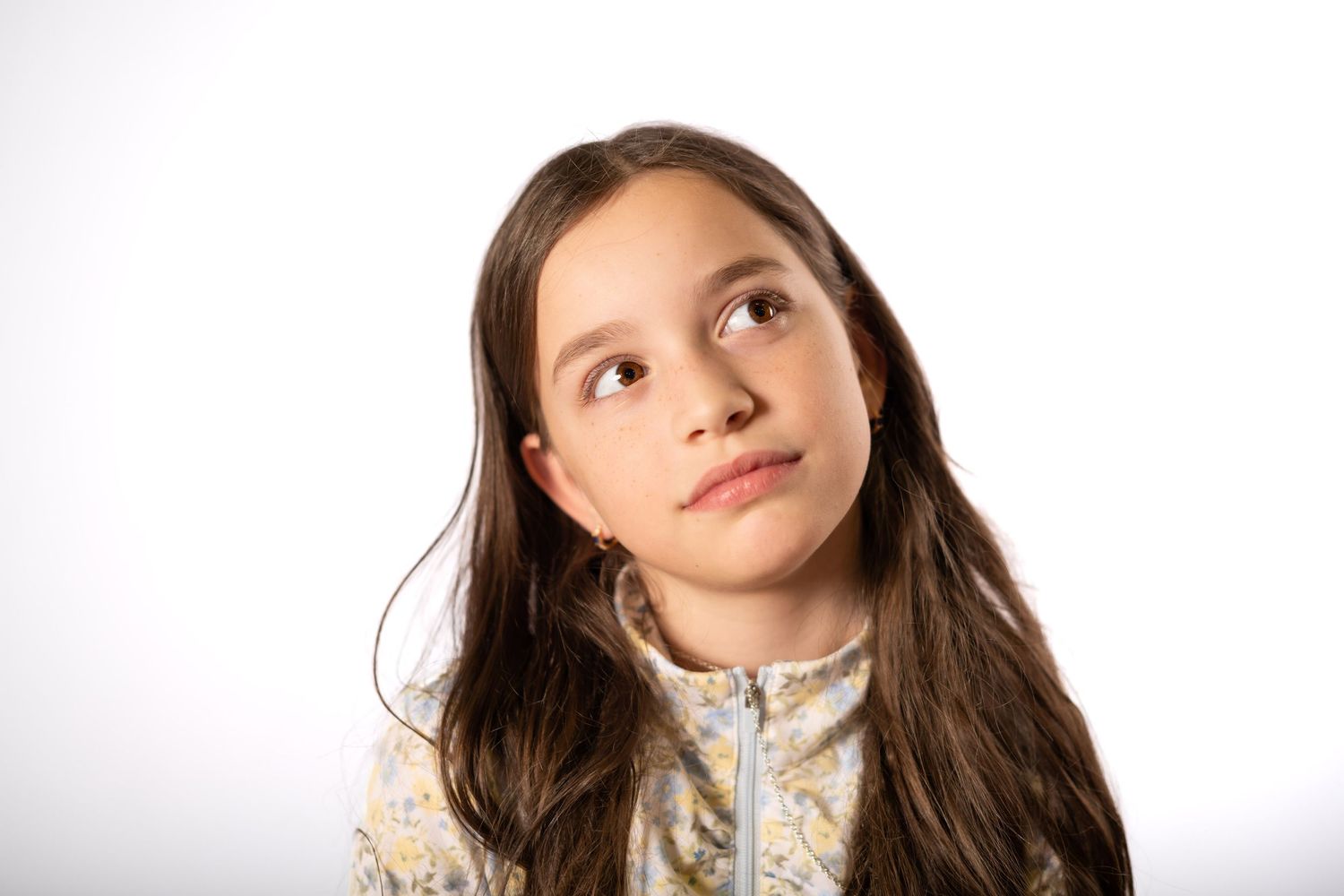 Young girl with long brown hair looking up thoughtfully against a white background.
