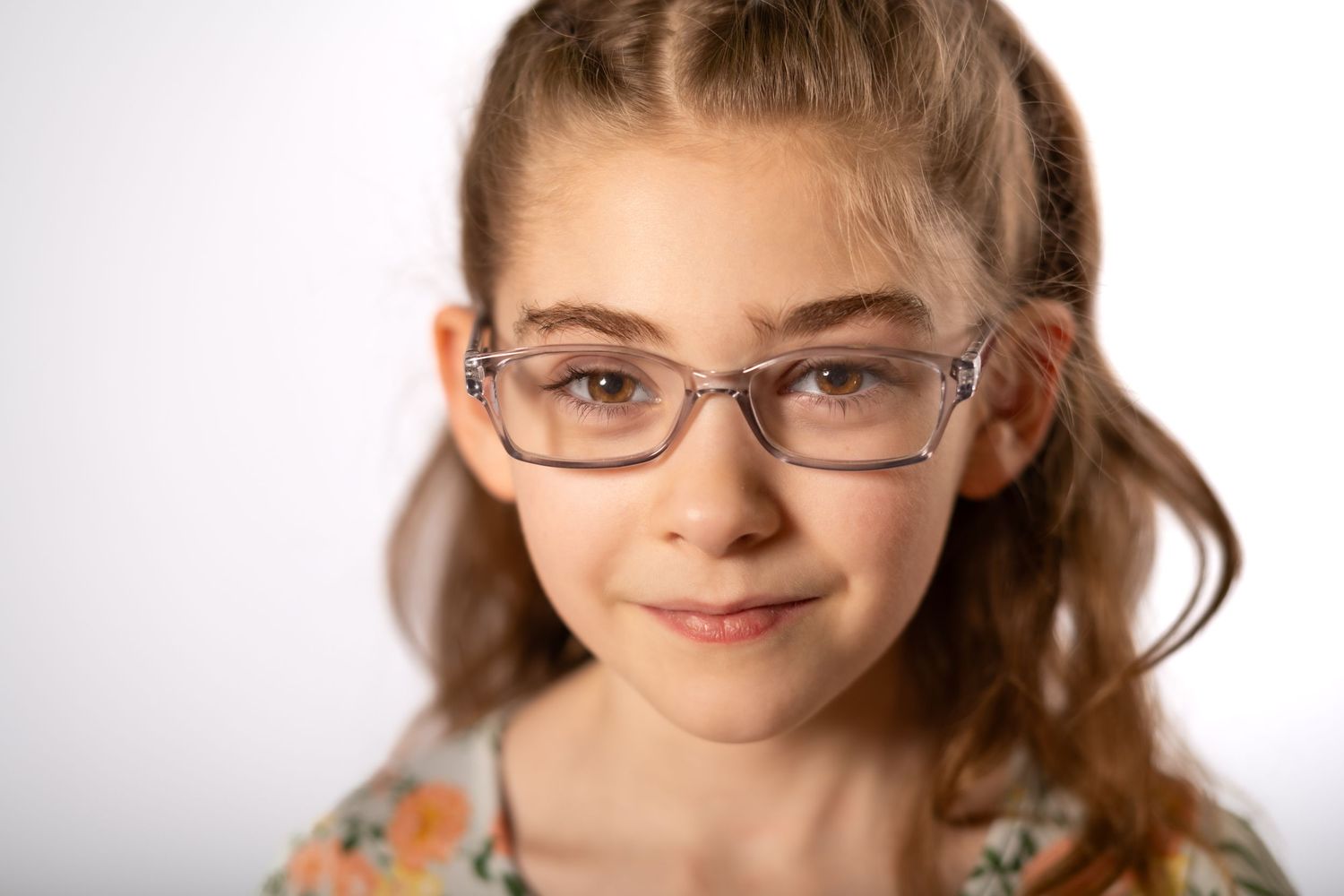Young girl with glasses and pigtails smiling at camera against white background.