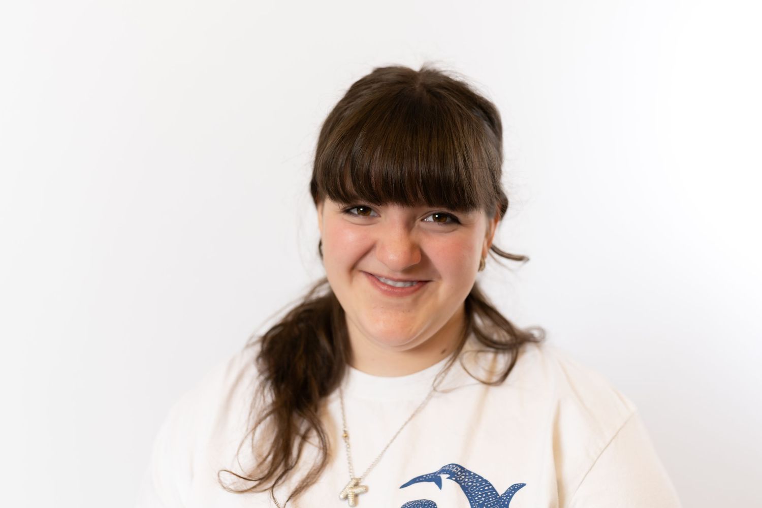 Young woman with dark hair and bangs smiling, wearing a white graphic tee with a necklace, against a white background.