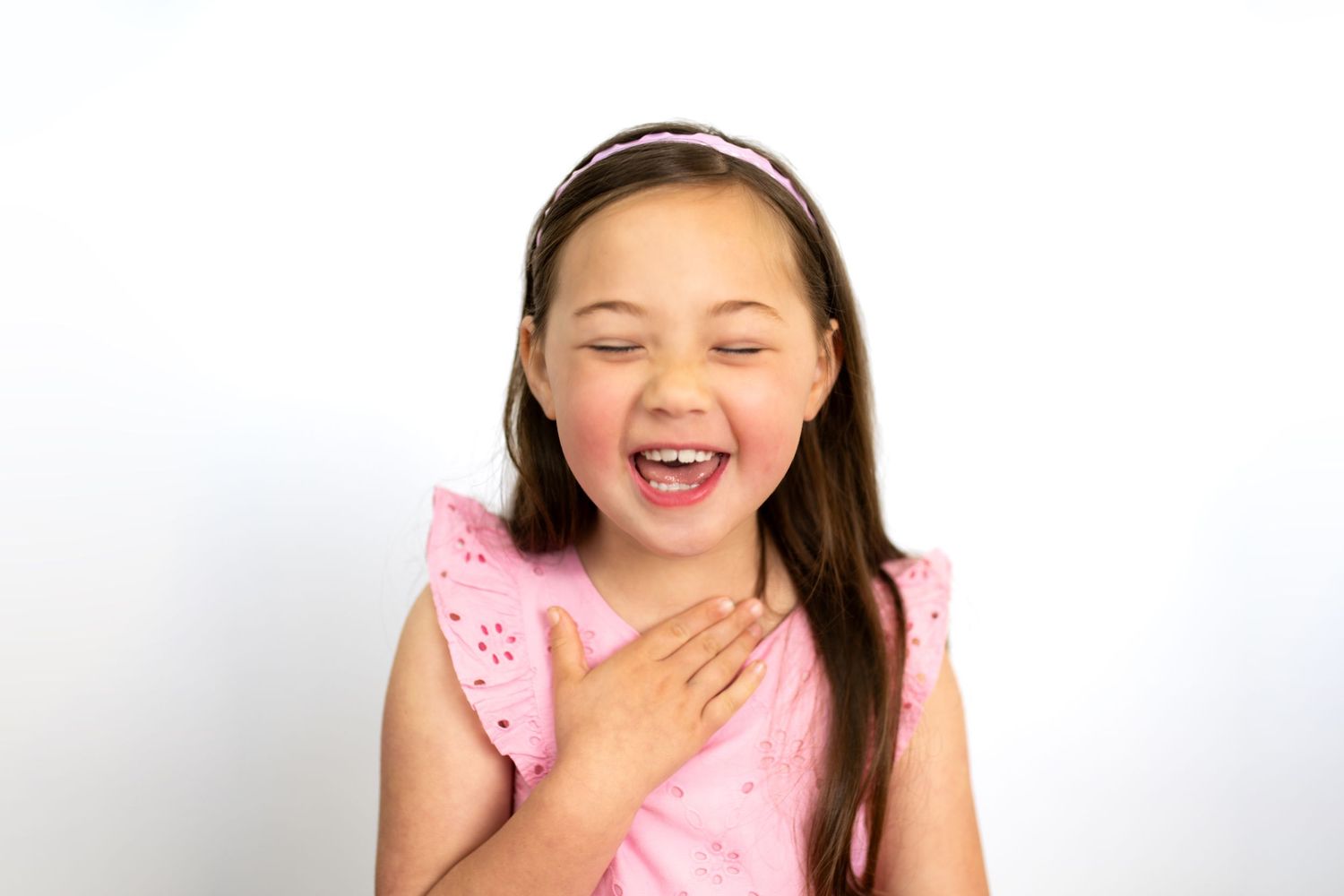 A young girl in a pink dress laughing joyfully with her hand on her chest against a white background.