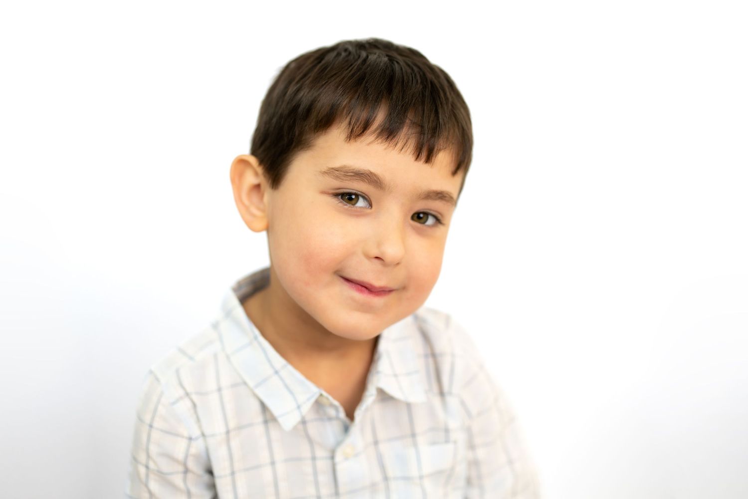 A young boy in a plaid shirt smiling softly against a white background in a portrait photo.
