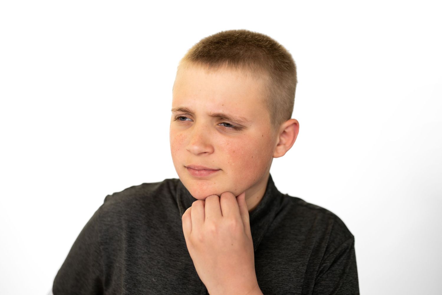 Thoughtful young boy with short hair touching his chin, wearing a dark gray shirt against a white background.