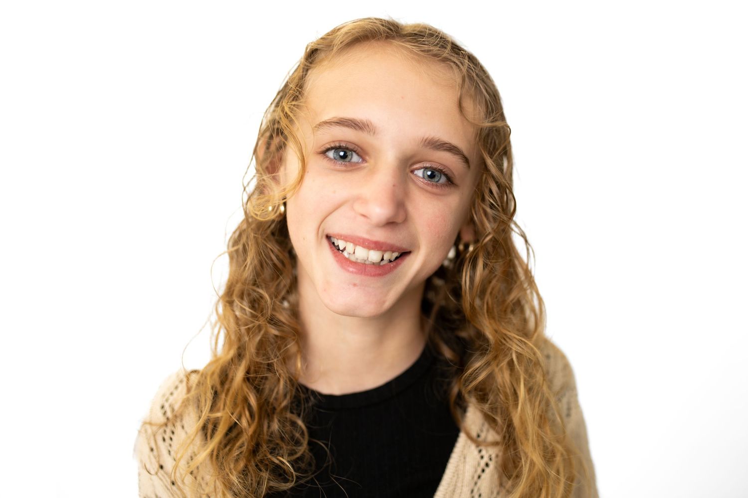 Smiling young girl with curly blonde hair and braces wearing a black top and beige jacket on white background.