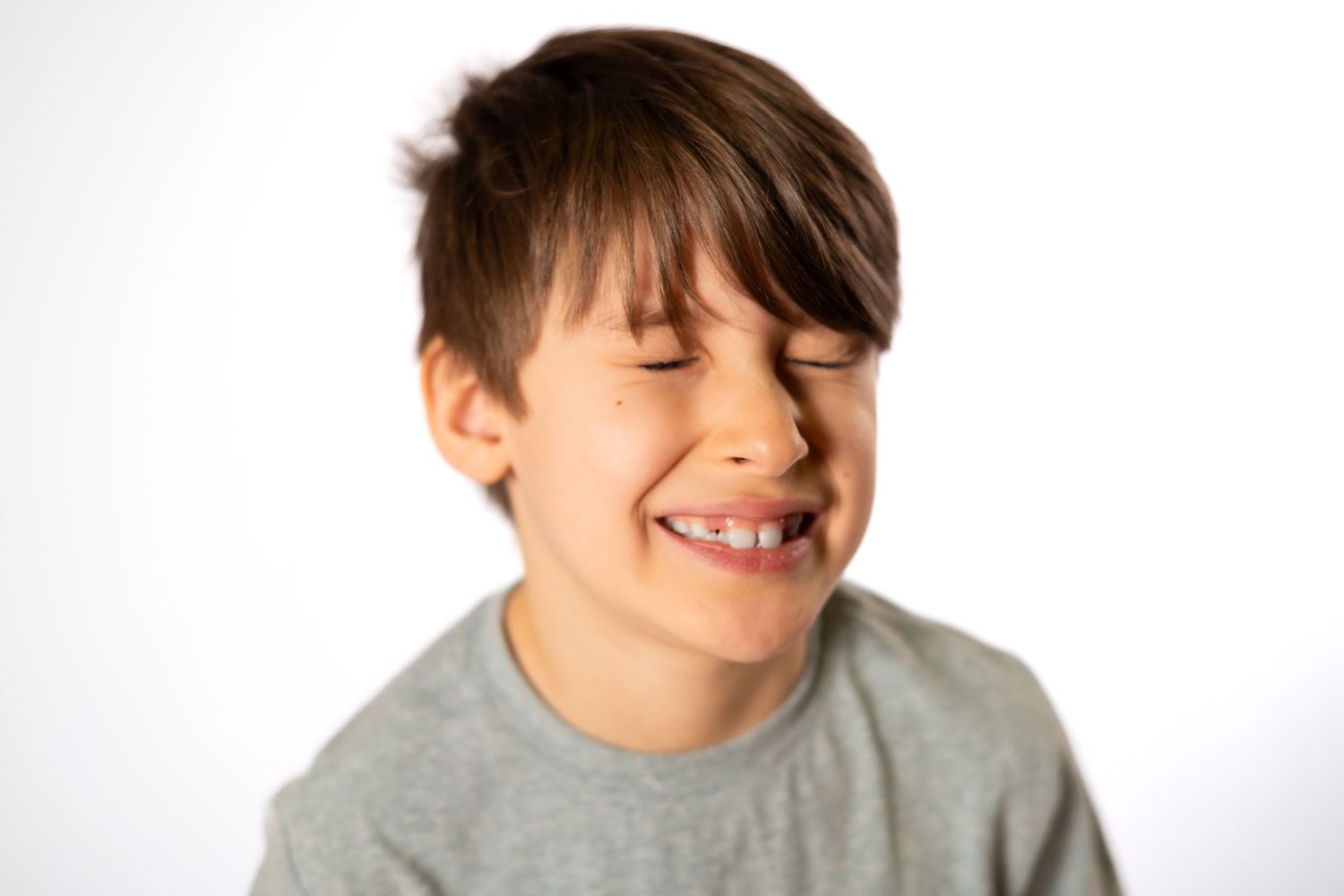Young boy with braces smiling with eyes closed, wearing a gray t-shirt against a white background.
