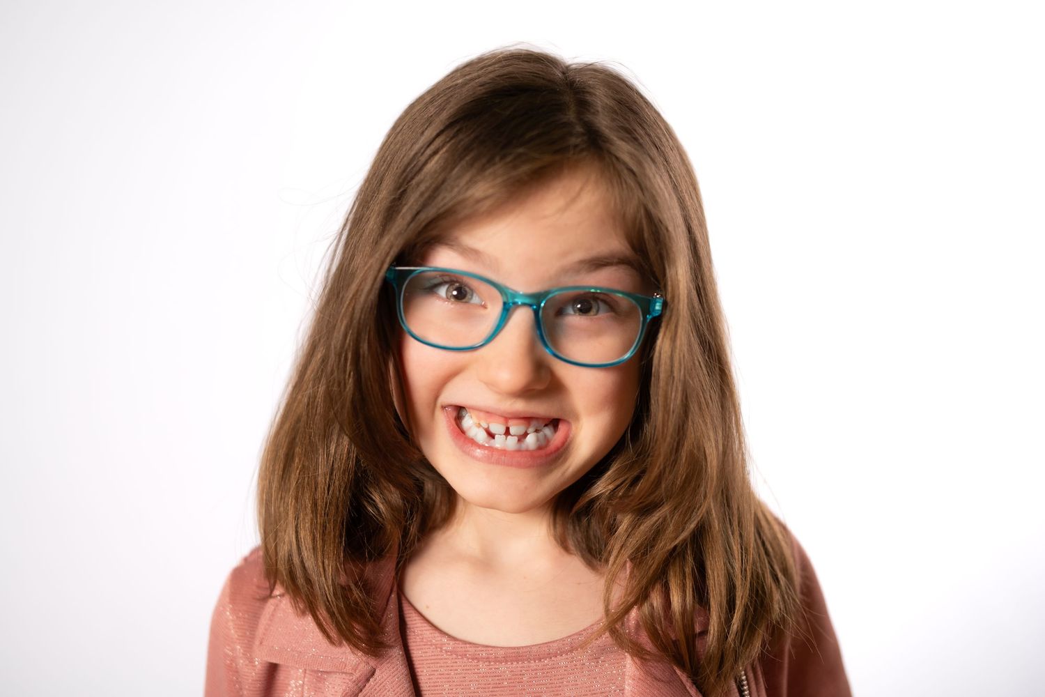 Young girl with teal glasses and long brown hair making a funny face against white background.