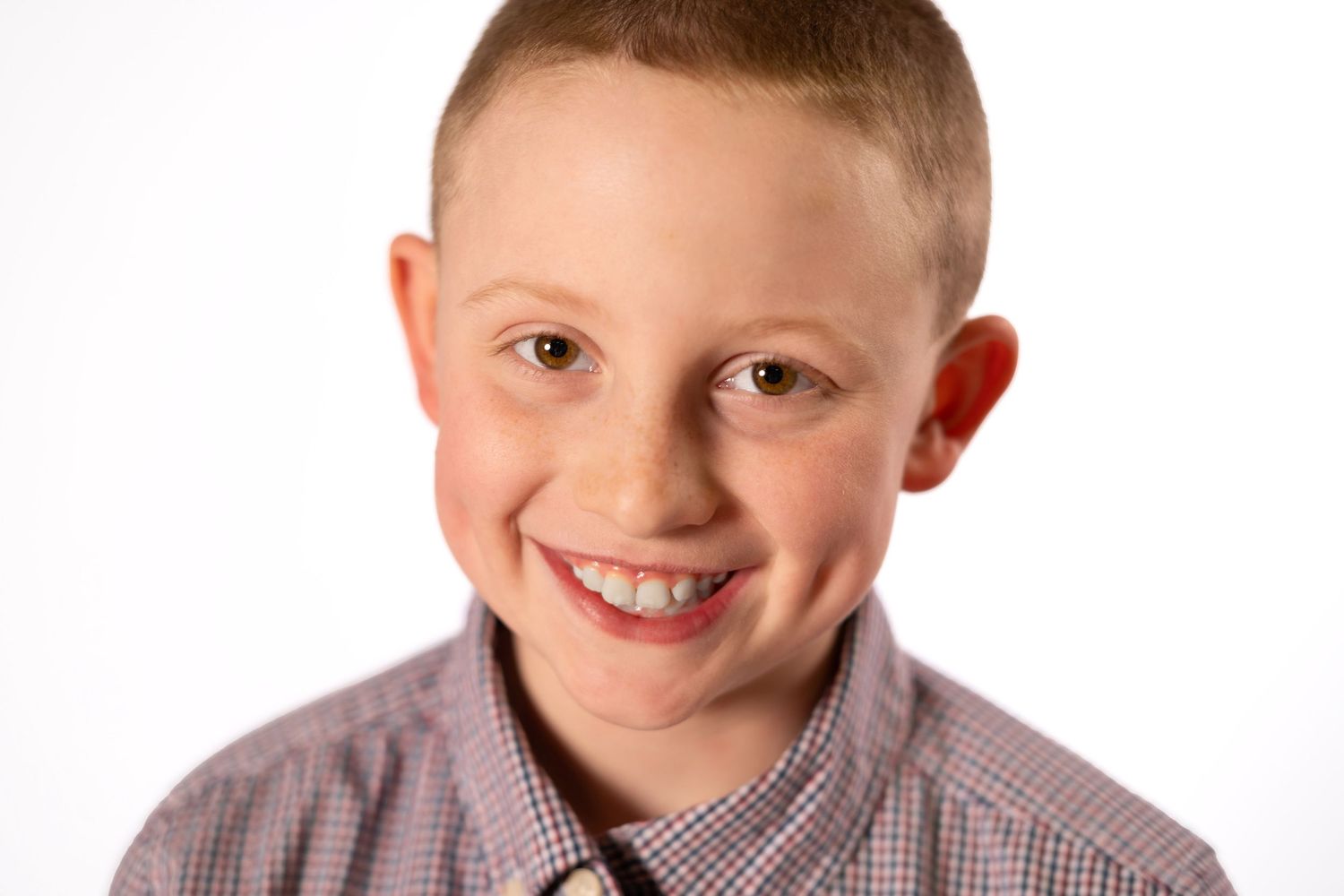 Smiling young boy with short hair wearing a plaid shirt, posing for a portrait photo against a white background.