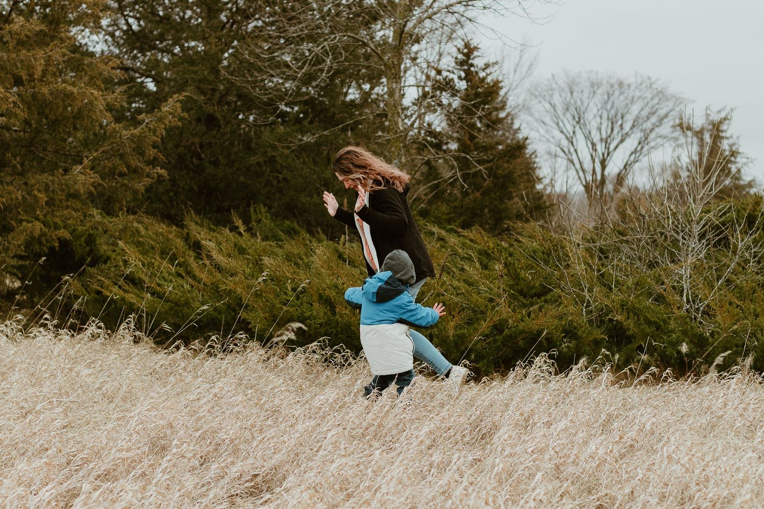 Two people embrace joyfully while standing in a tall grass field with evergreen trees in the background.