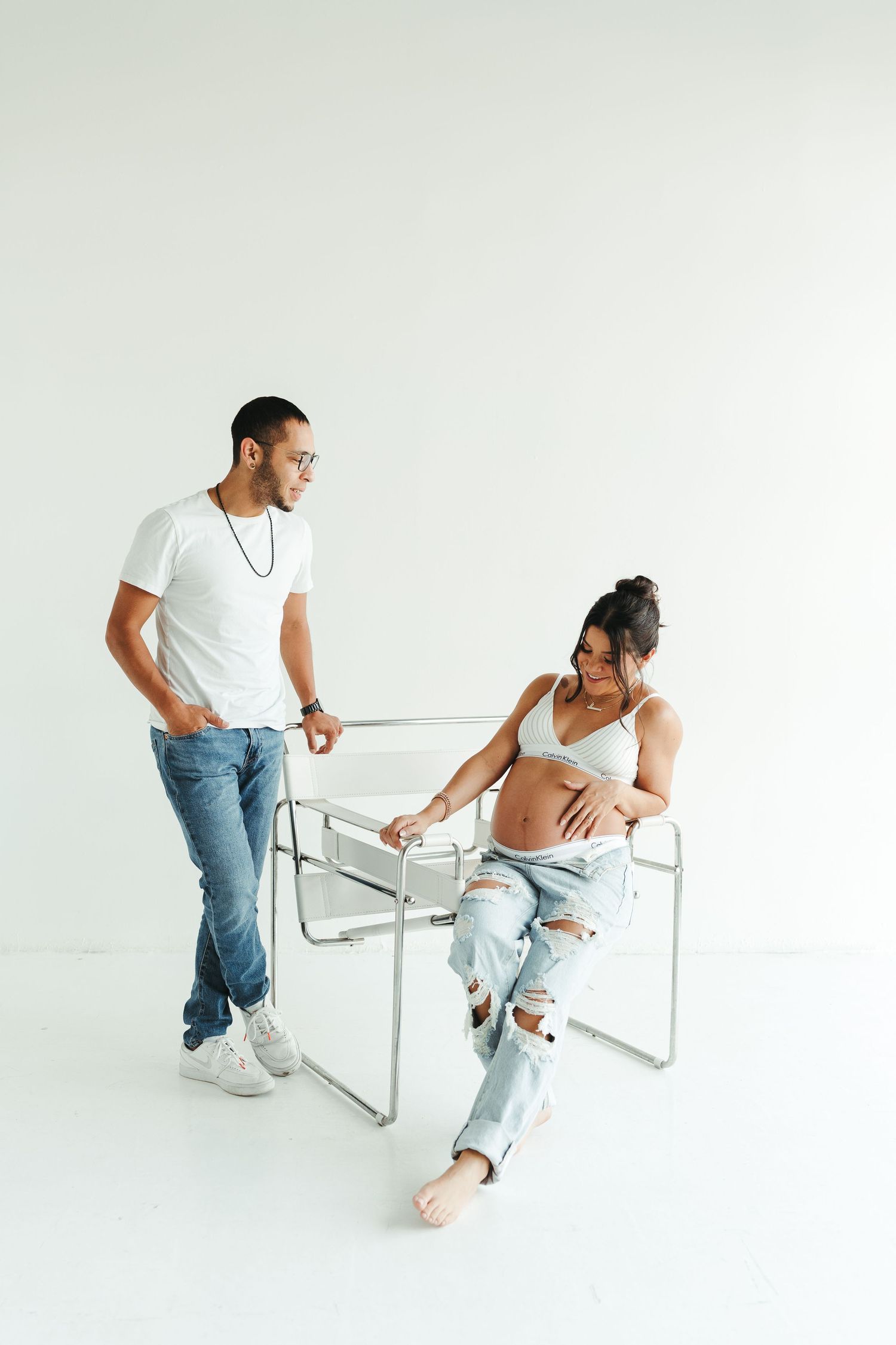 Couple in casual white outfits interacting playfully with a modern white chair against a minimalist background.