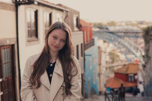 Woman in beige coat standing against urban backdrop.