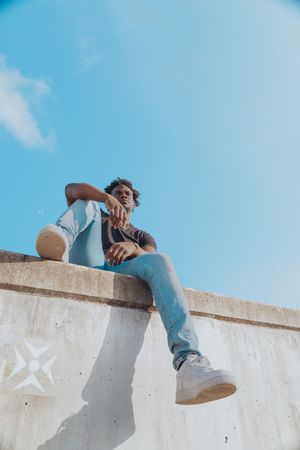 Person in casual wear sitting on concrete wall against blue sky.