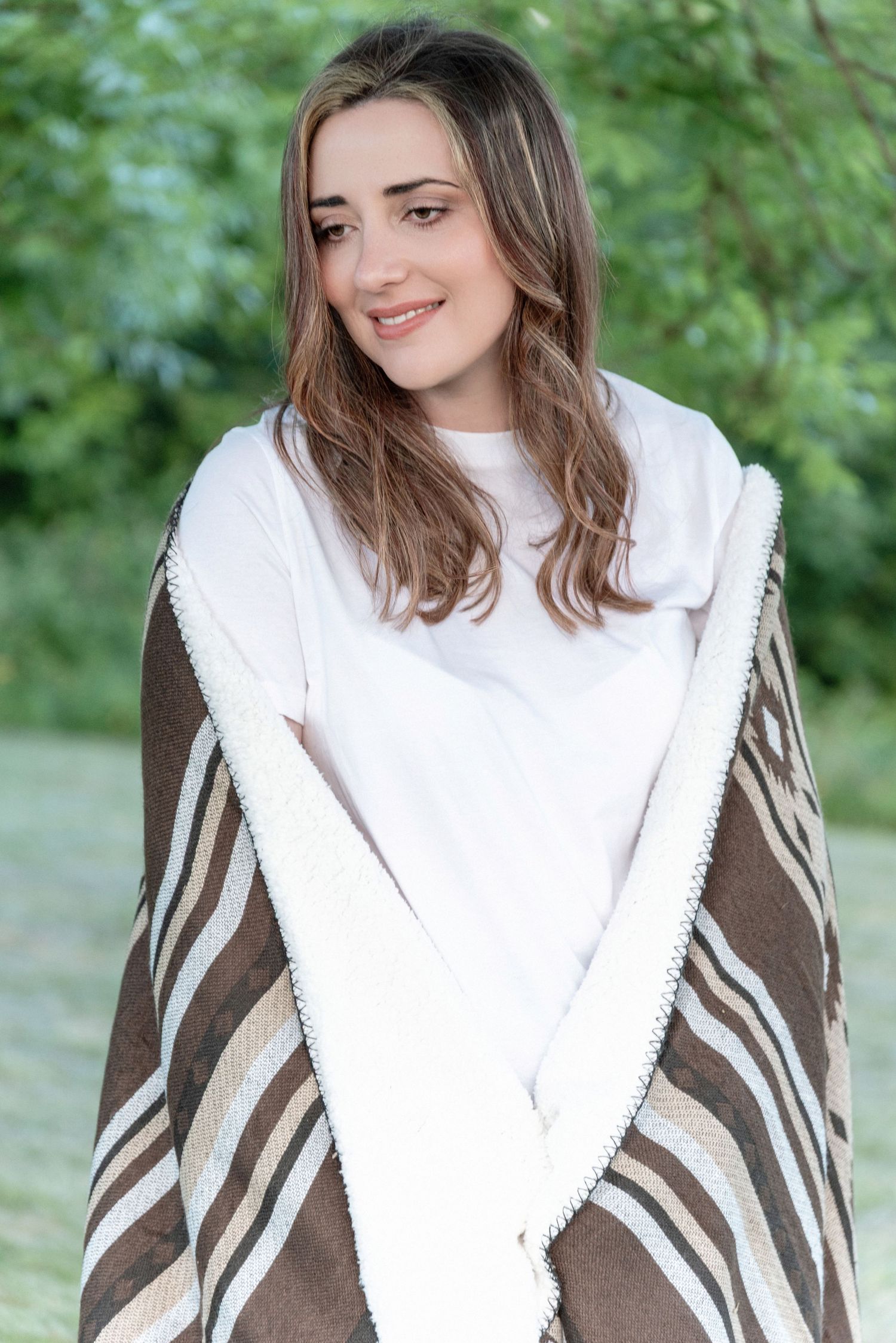 Someone wrapped in a cozy brown and white striped blanket smiles while wearing a white sweater outdoors.