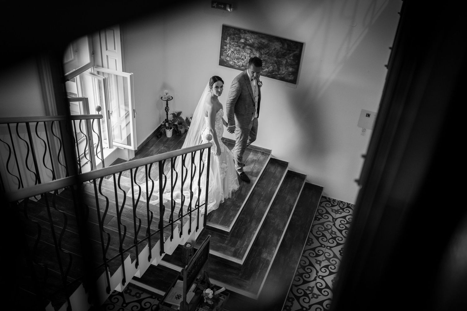 A black and white photograph shows a bridal gown against an ornate staircase railing in an elegant interior setting.